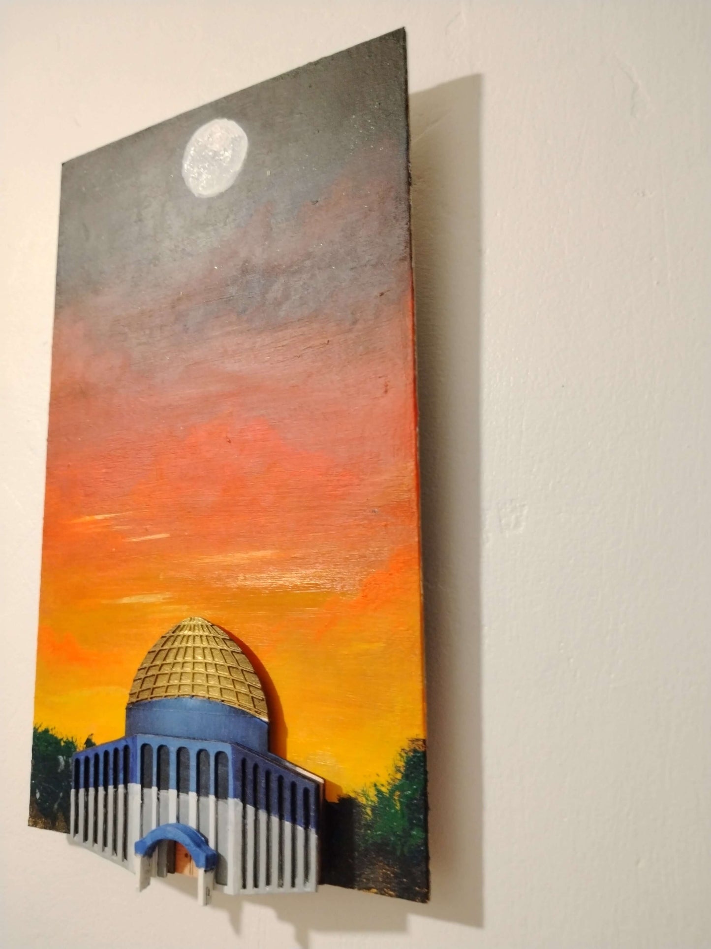 A vertical hand-painted wooden wall art piece depicting a domed classical building under a sunset sky with a moon, mounted on a white wall.