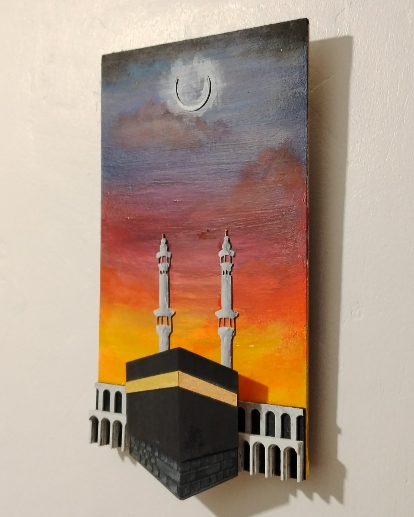 A vertical rectangular wall art piece depicting a mosque with two minarets against a sunset sky, with a crescent moon near the top. The artwork is mounted on a plain light wall and casts a soft shadow to the right.