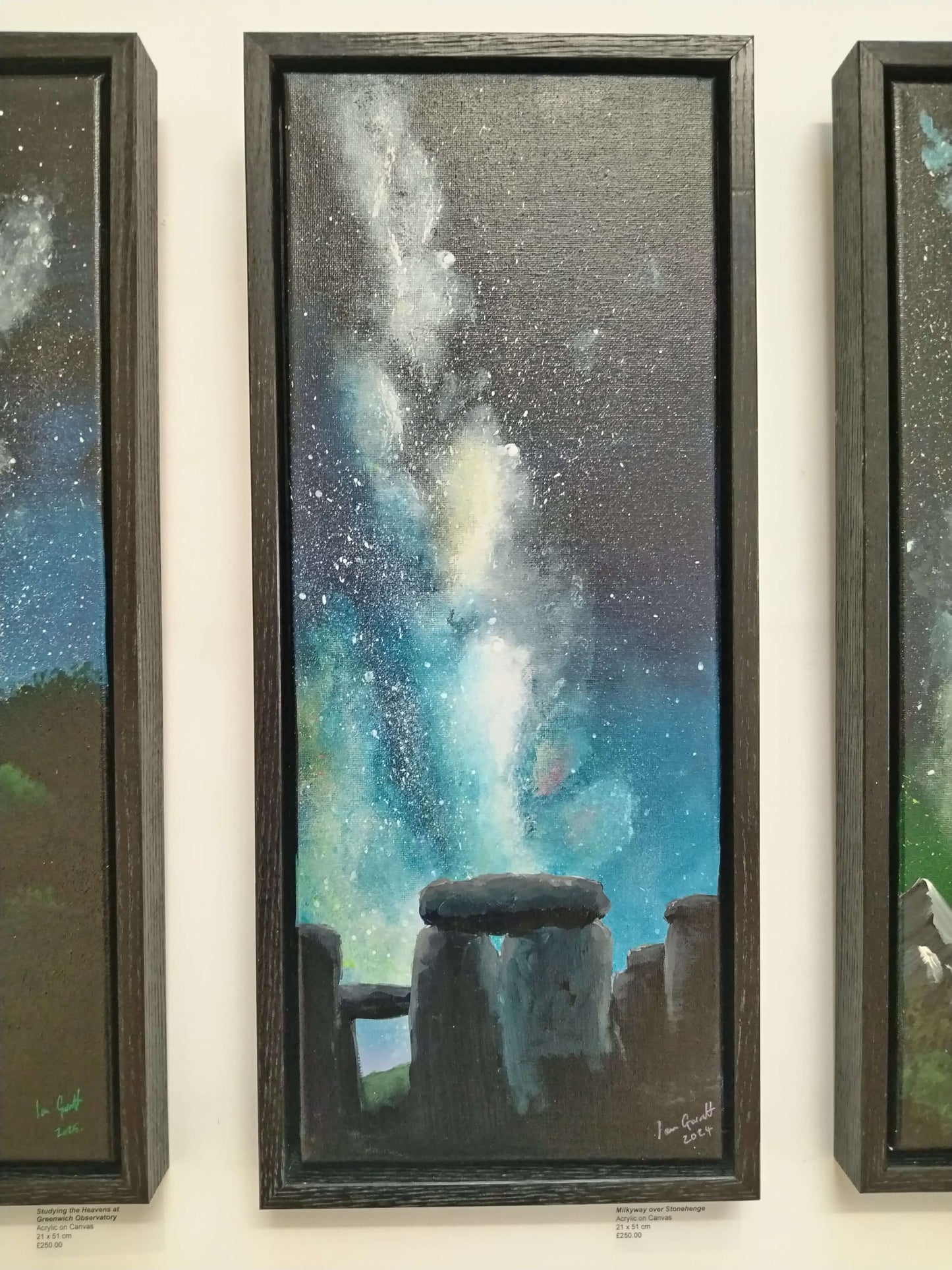 A framed painting of the Milky Way rising over the standing stones of Stonehenge hanging on a white wall.