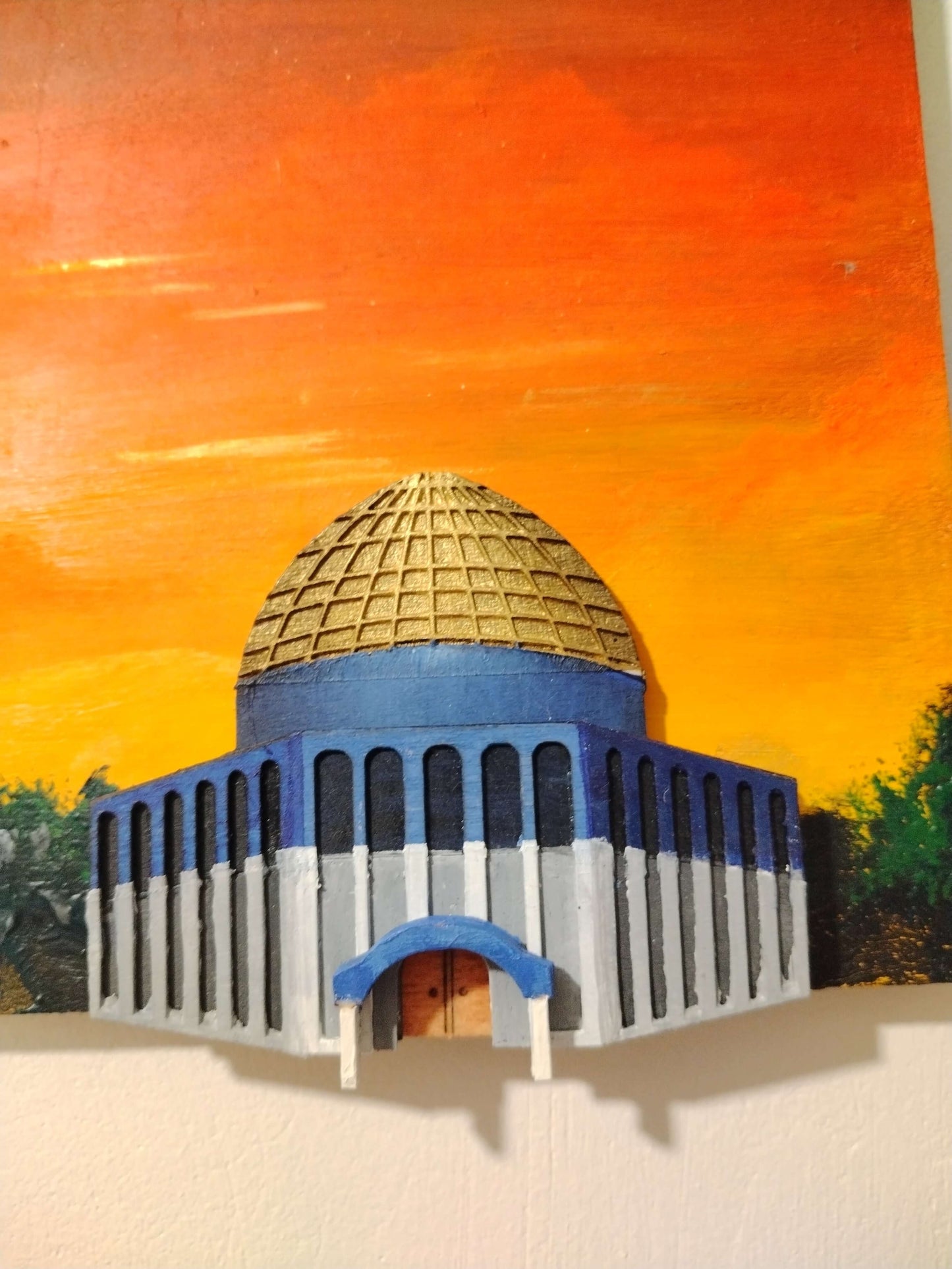 A miniature painted model of a domed building, likely a mosque or historic structure, featuring a blue and white facade with a gold dome, set against a vibrant orange and yellow sunset sky.