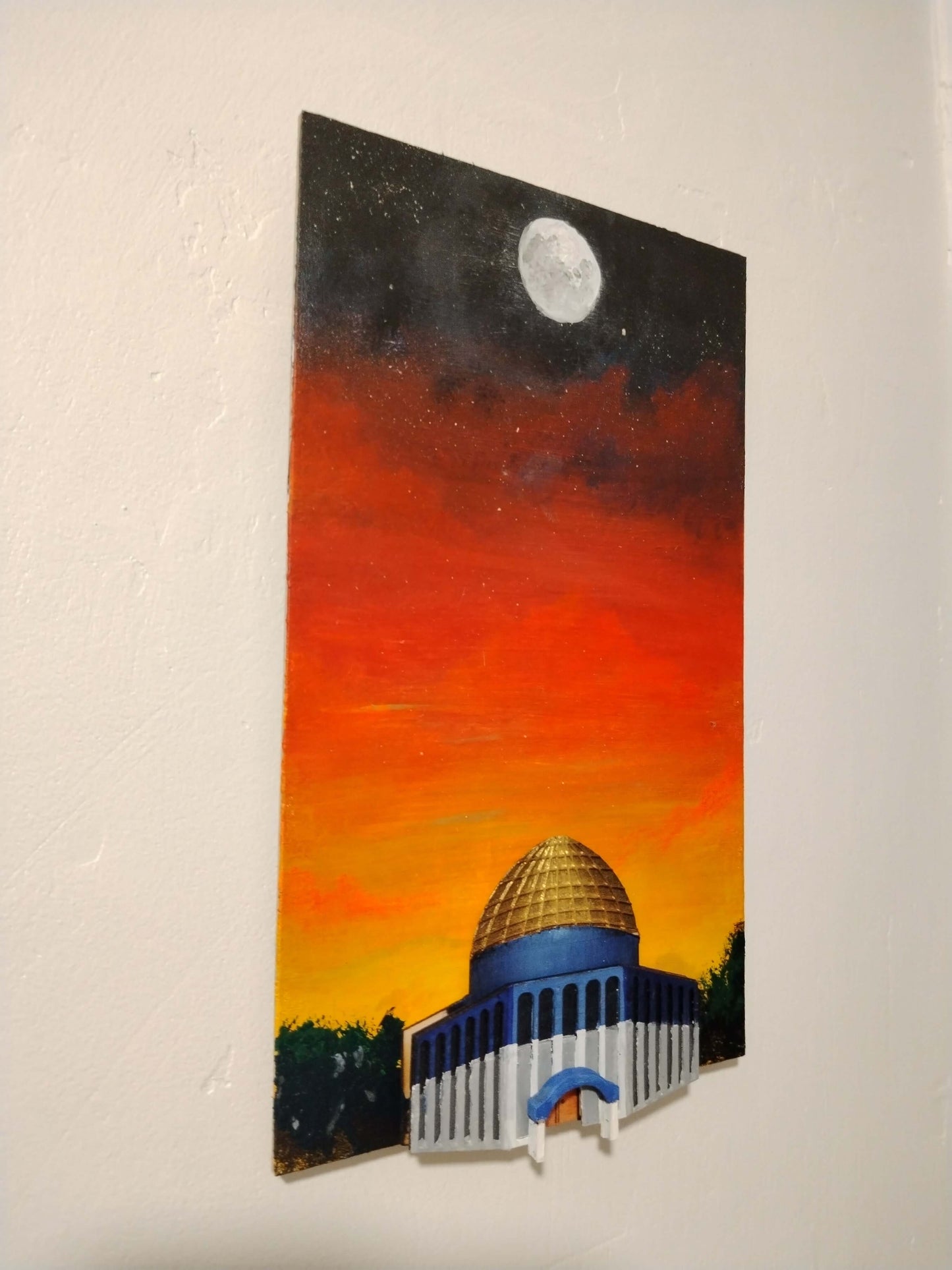 A vertical canvas painting of a domed building under a starry night sky with a bright moon, painted in orange, red, black, blue, and white, mounted on a textured off-white wall.