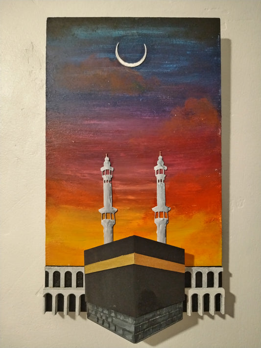A vertical hand-painted 3D wooden wall art piece depicting the Kaaba and two minarets under a crescent moon against a sunset sky gradient of blue, purple, orange, and red, mounted on a beige wall.