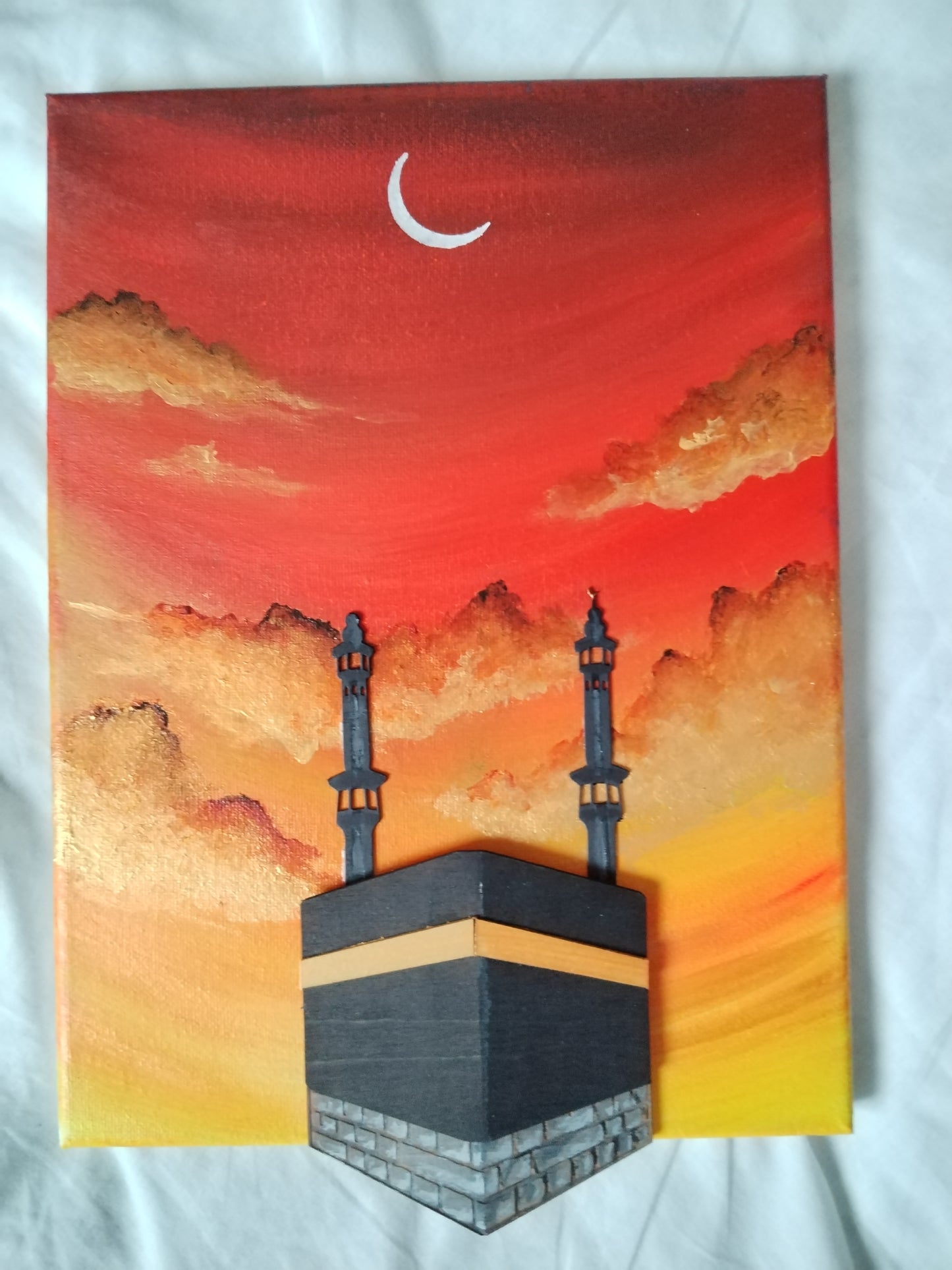 A rectangular canvas painting depicting the Kaaba at sunset, with a red and orange sky, clouds, a white crescent moon, and two minarets, photographed on a white fabric background.