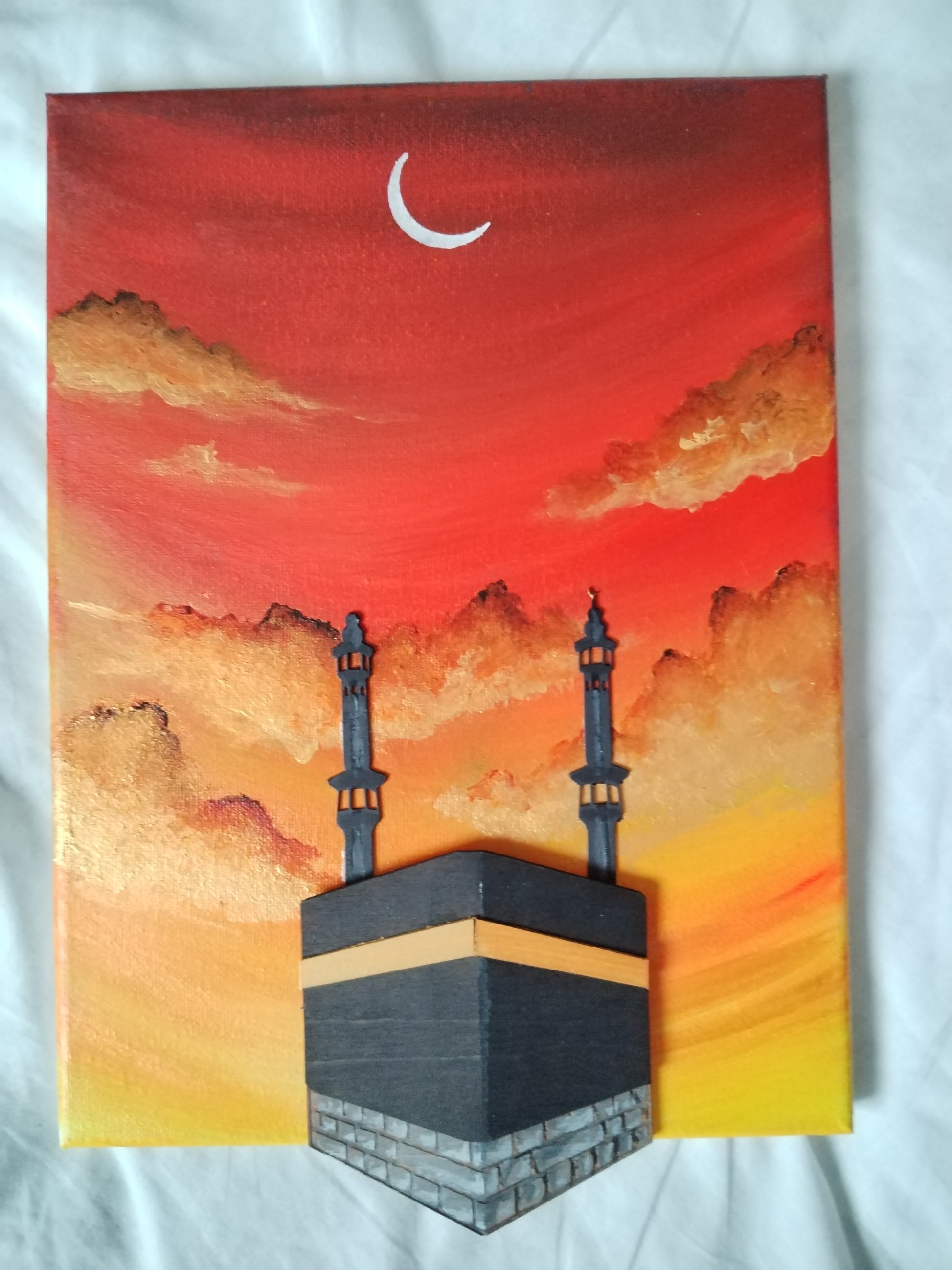 A rectangular canvas painting depicting the Kaaba at sunset, with a red and orange sky, clouds, a white crescent moon, and two minarets, photographed on a white fabric background.